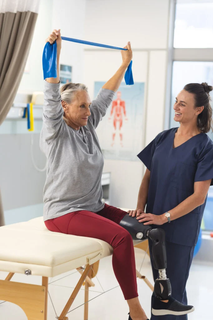 Caucasian female physiotherapist and senior woman with artificial leg using exercise band stretching. Hospital, disability, physiotherapy, work, medicine and healthcare, unaltered.