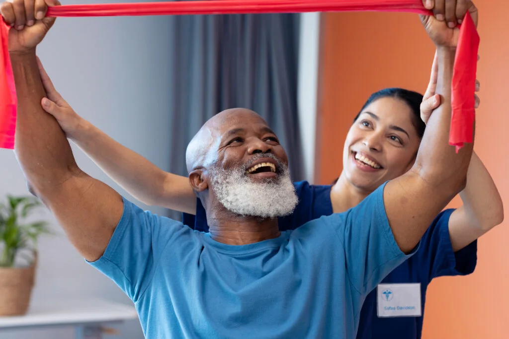 Smiling diverse female physiotherapist and senior male patient exercising with band. Hospital, medical and healthcare services.