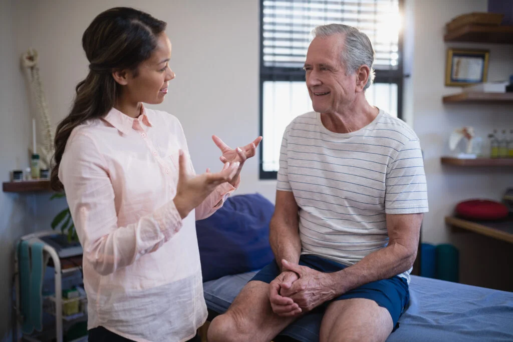 Senior male patient looking at female therapist gesturing while talking in hospital ward